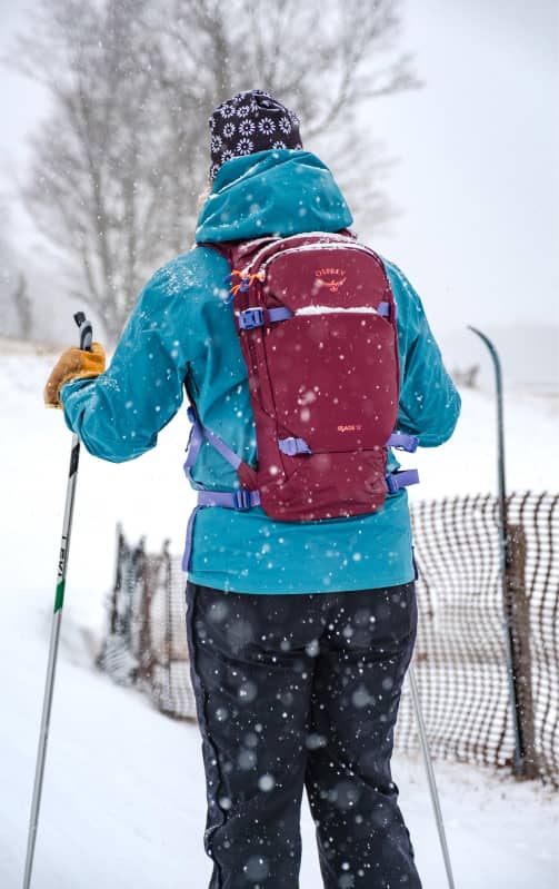 Cross‑country skier moving through snowy terrain while wearing an Osprey backpack, with falling snow and winter landscape in the background.
