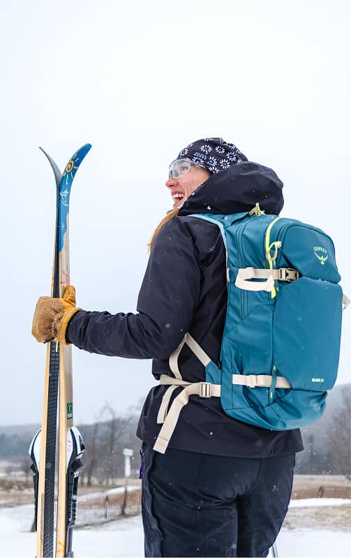 Cross‑country skier holding skis while wearing an Osprey backpack in a snowy winter landscape.