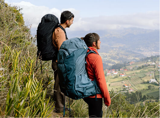 Two hikers carrying large Osprey backpacks standing on a mountainside overlooking a rural valley.
