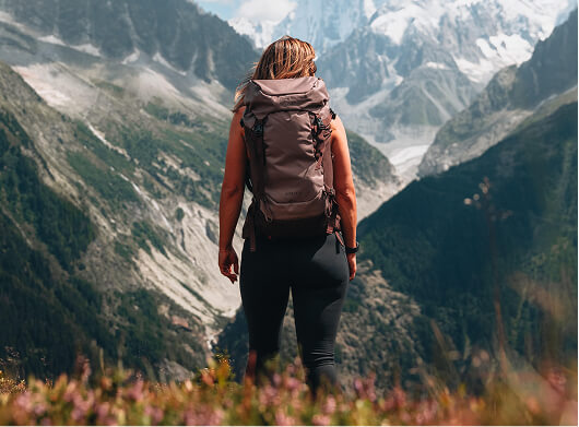 Hiker wearing a backpack standing on a mountain trail overlooking a rugged alpine valley.