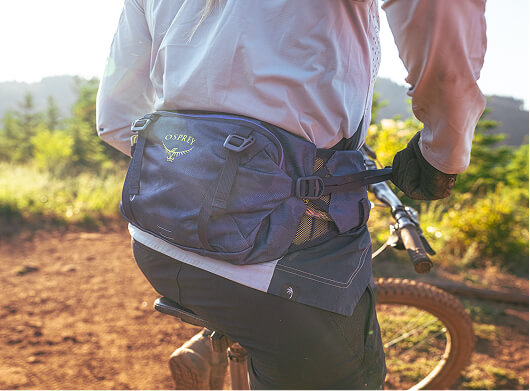 Cyclist wearing an Osprey waist pack riding a mountain bike on a sunlit dirt trail.