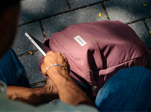 Person placing a laptop into an Osprey backpack while sitting on an outdoor paved area.
