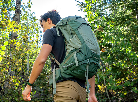 Hiker wearing an Osprey backpack walking through a sunlit forest trail.