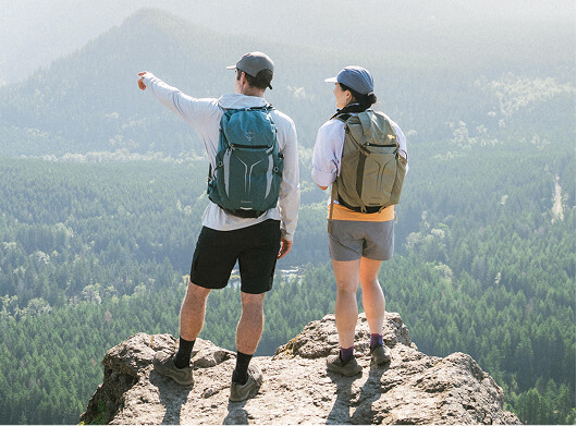 Two hikers wearing Osprey backpacks standing on a rocky overlook pointing out across a forested valley.