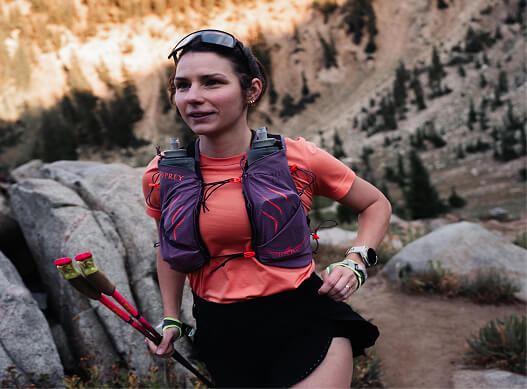 Trail runner wearing an Osprey running vest moving along a rocky mountain path with trekking poles.
