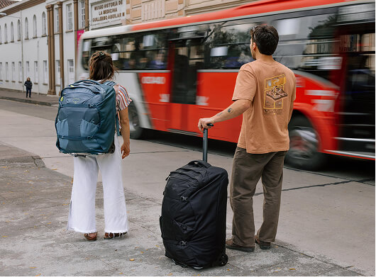 Travelers with Osprey luggage waiting at a city bus stop with rolling suitcases and a large travel pack.