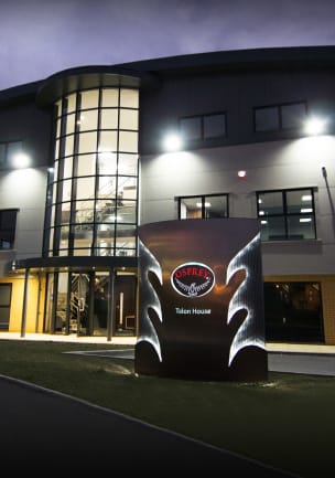 Et nattbilde av et treetasjes kontorbygg med buet tak og glassatrium i flere etasjer ved hovedinngangen. På en gressflekk foran bygget står det et stort, opplyst skilt med Osprey-logoen. Utskjæringer i form av vinger og navnet «Talon House» er opplyst.