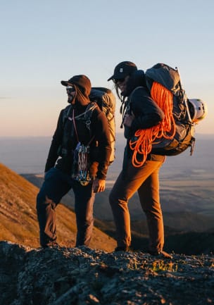 To klatrere står i morgenlys på en fjellrygg med hver sin fullpakkede ryggsekk på ryggen, med klatretau, karabinkroker, hjelmer og isøks.