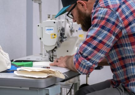En skjeggete mann i briller, caps og rød, blå og hvit flanellskjorte med oppbrettede ermer syr et munnbind i stoff på en symaskin.