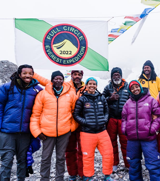 Et team på syv klatrere i fjellklatrebukser og dunjakker under et hvitt flagg med grønn stripe og et blått, rødt og gult emblem med påskriften «Full Circle Everest Expedition 2022», og tibetanske bønneflagg som blåser i vinden bak dem.