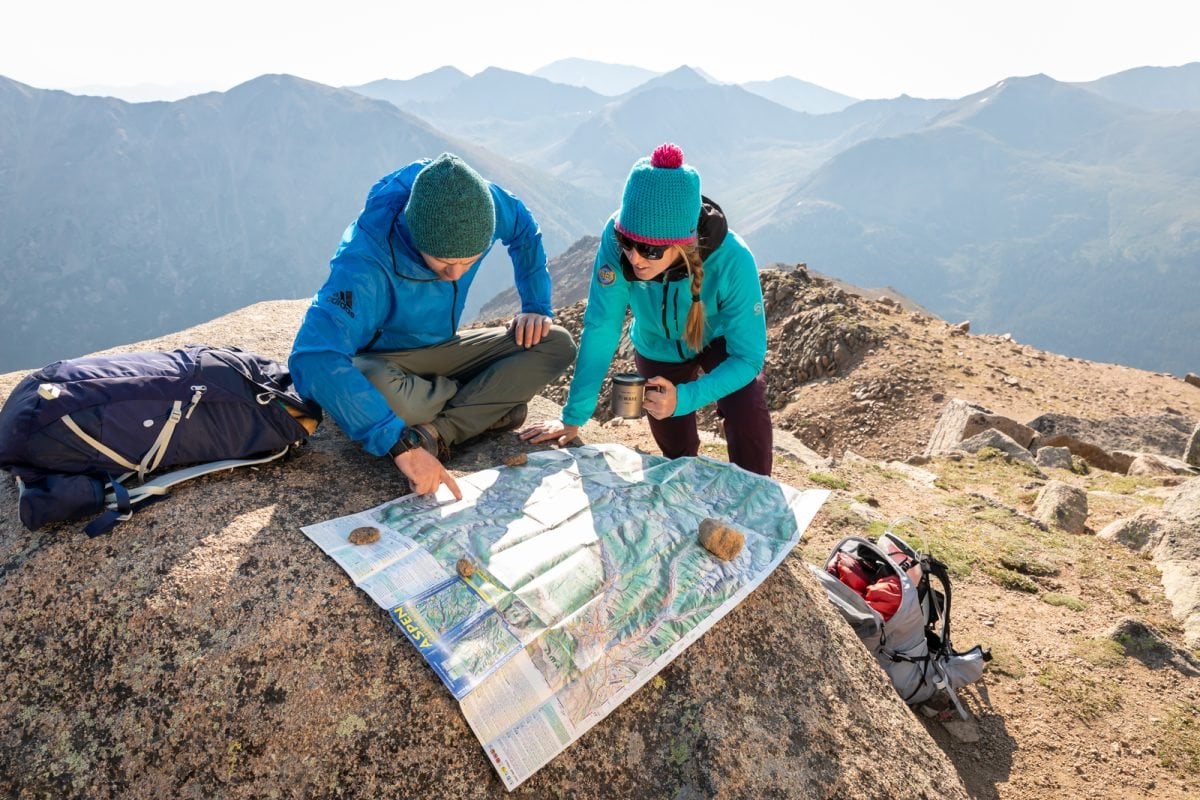 A man and a women looking at a map that is laid over over a rock on top of a mountain.