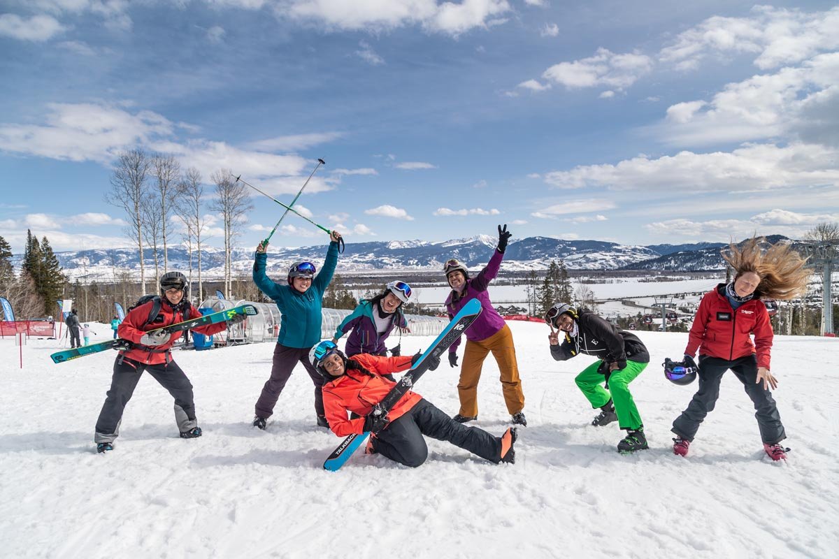 Seven women wearing ski gear or holding skis, posing for a silly picture
