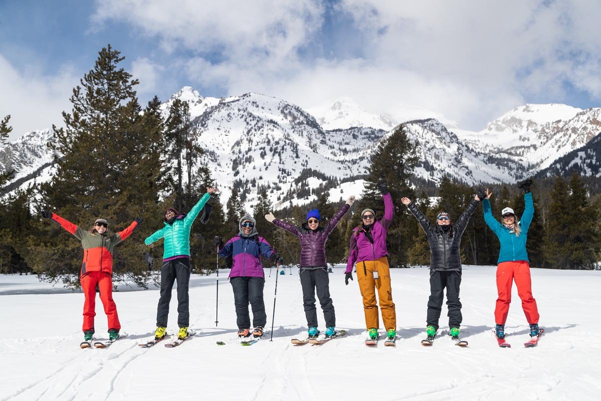 Seven woman wearing skis on a snowy mountain posing with their hands in the air