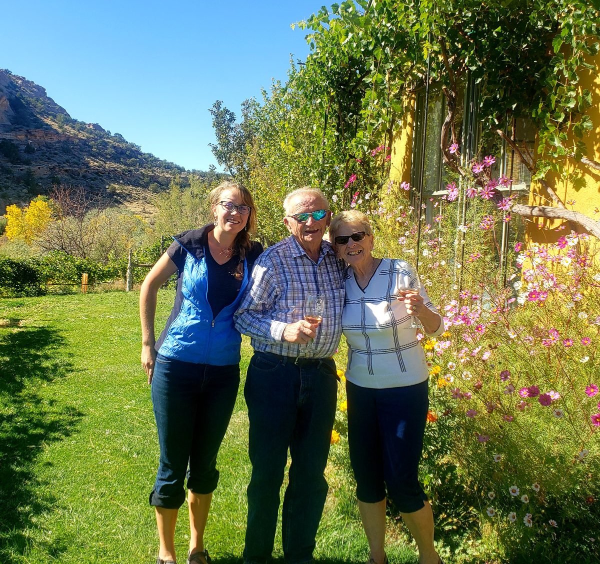 Three people standing together, two holding glasses of wine, with a yellow house and grass in the background