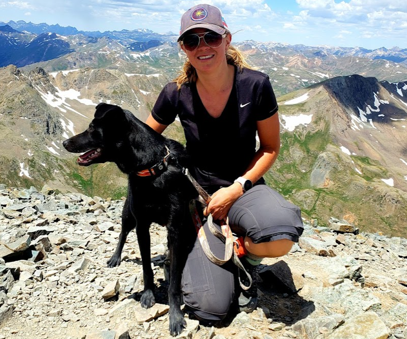 A woman with her black dog posing at the top of our mountain, with mountain ranges in the background