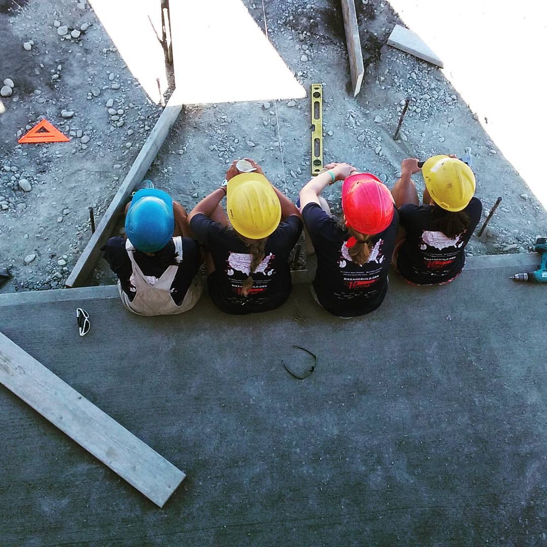 Four people sitting on a concrete ledge, wearing different colored hard hats