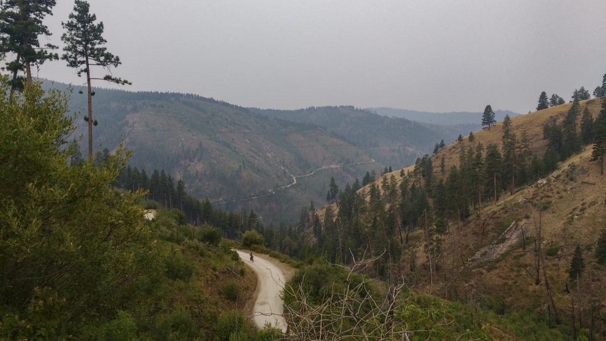 A landscape view of a biker riding down a dirt road, with mountains on either side.