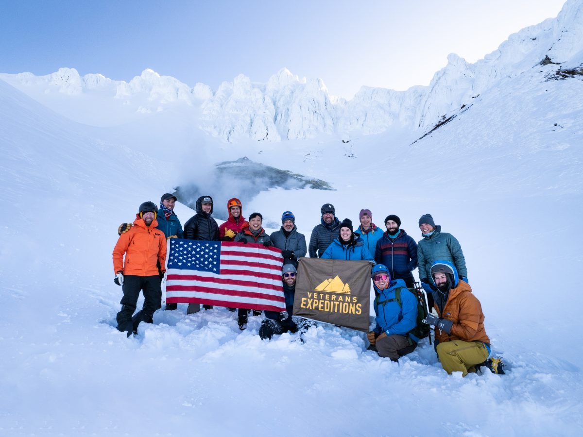 A group of people standing atop a snowy mountain, holding an American flag and a Veterans Expeditions flag.