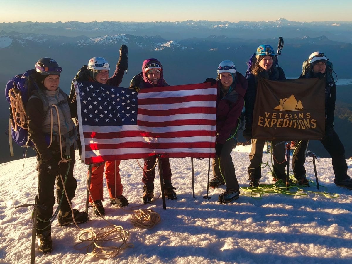 A group of people standing atop a snow mountain holding an American flag and a Veterans Expedition flag.