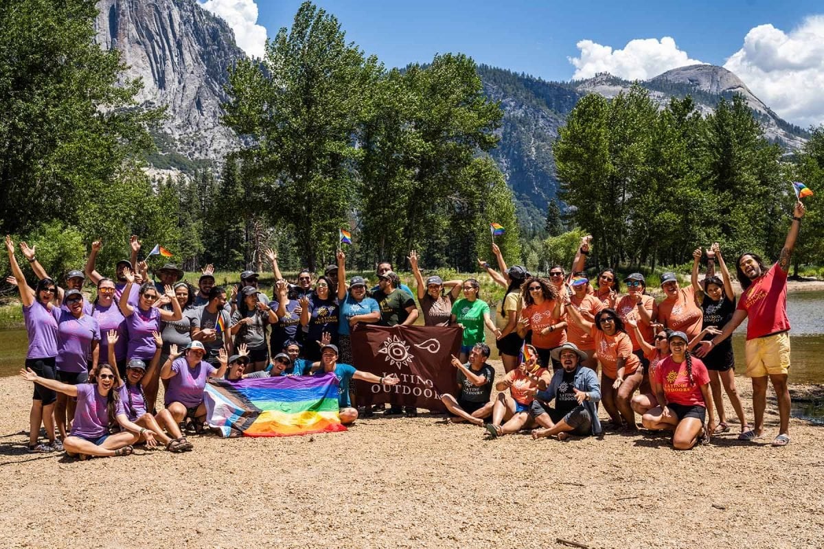 A large group of people near the water's edge posing and holding a Latino Outdoors flag and an LQBTQIA+ flag
