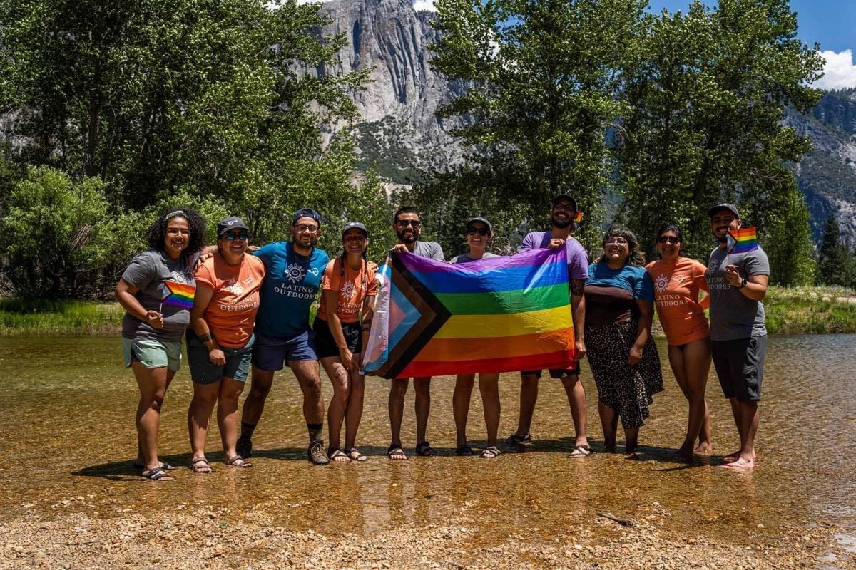 A group of people standing in very shallow water holding a large LQBTQIA+ flag, and two people holding small flags