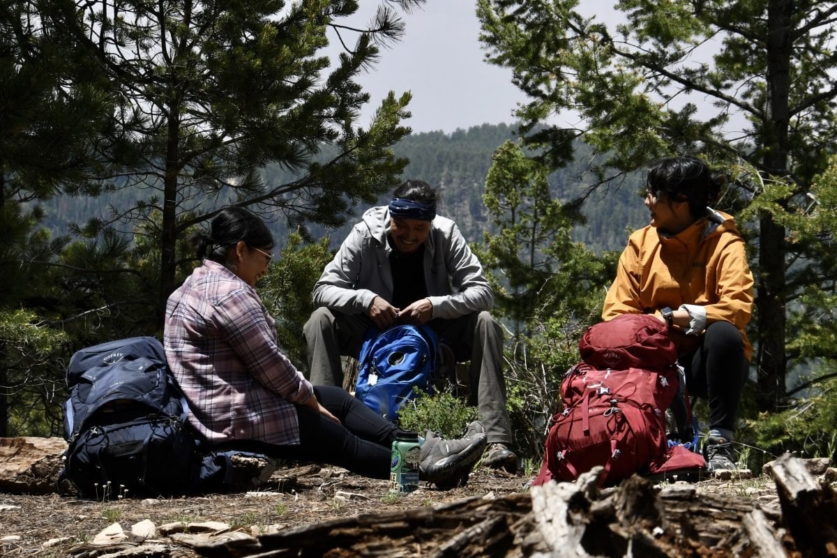 Three people sitting ono the ground in the forest, with backpacks next to them