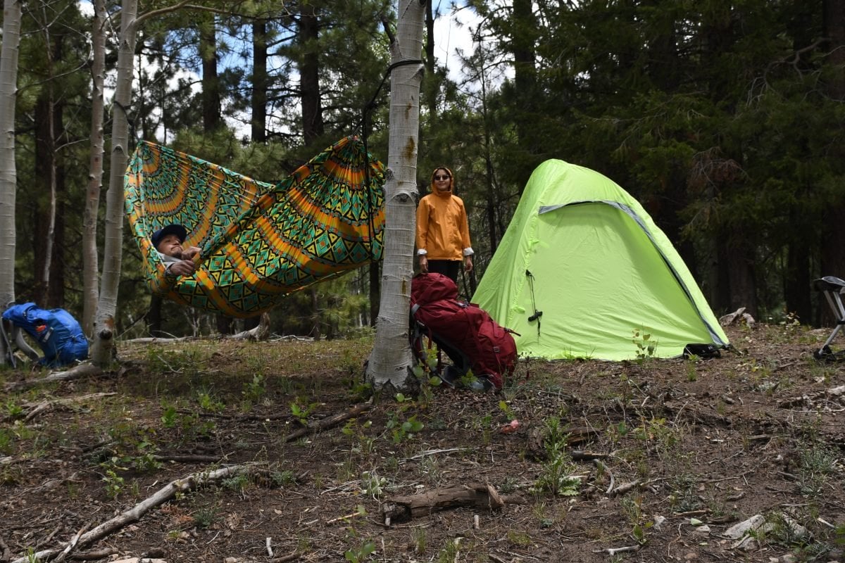 A person hanging in a green and orange hammock, with another person standing in the background next to a green tent