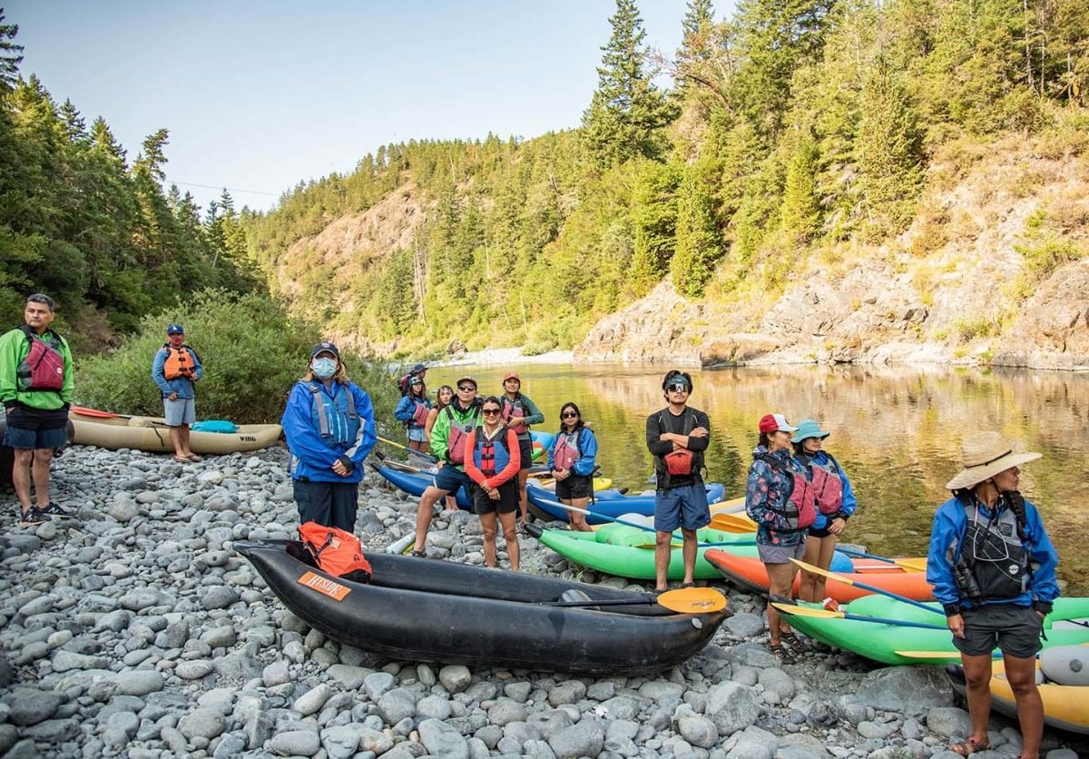 A group of people standing on a riverbank of rocks wearing kayaking gear and life jackets, with kayaks on the ground around them