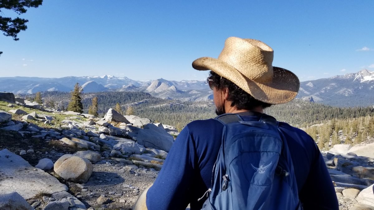A man facing a mountain range in the distance, wearing a tan hat and a blue backpack.