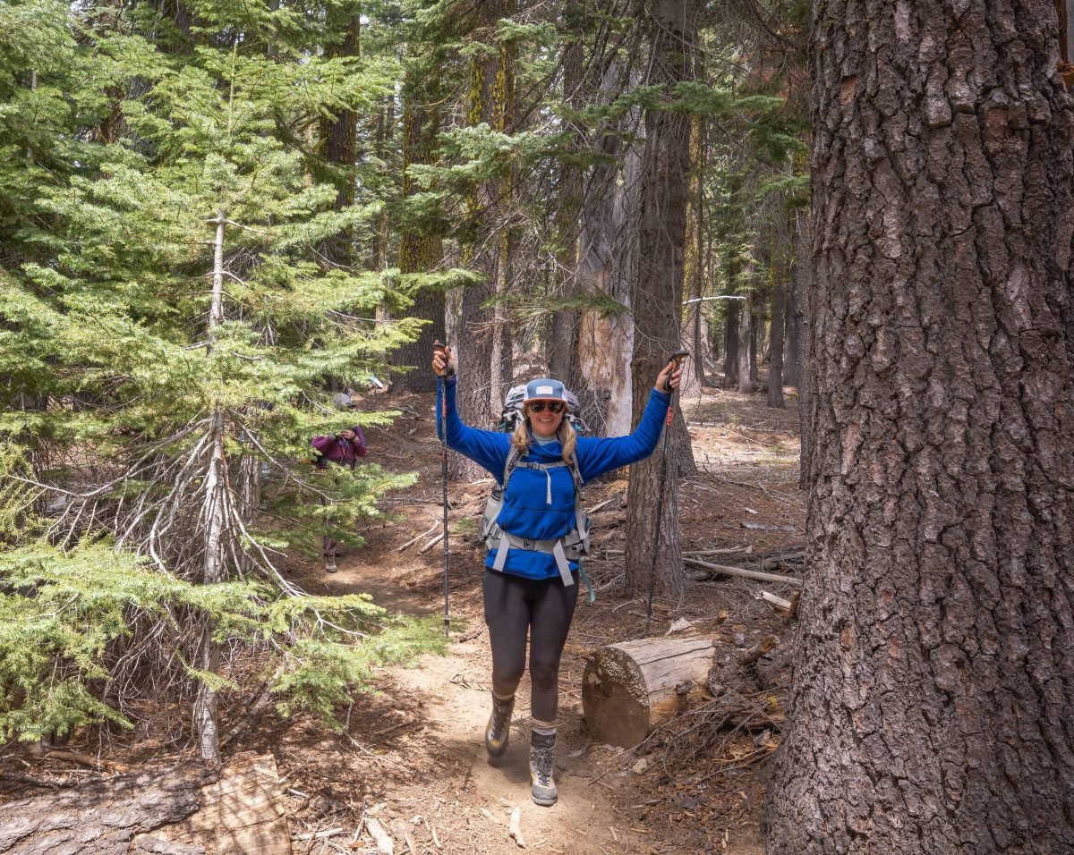 A woman geared up with mountaineering gear holding trekking poles up along a forested trail
