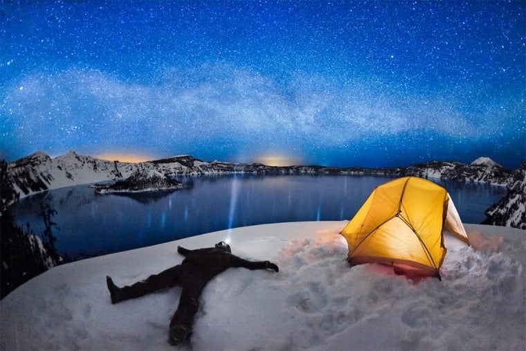 A person laying in the snow facing the sky, wearing a headlamp, with a sky full of stars above, and a lit, yellow tent to the side.