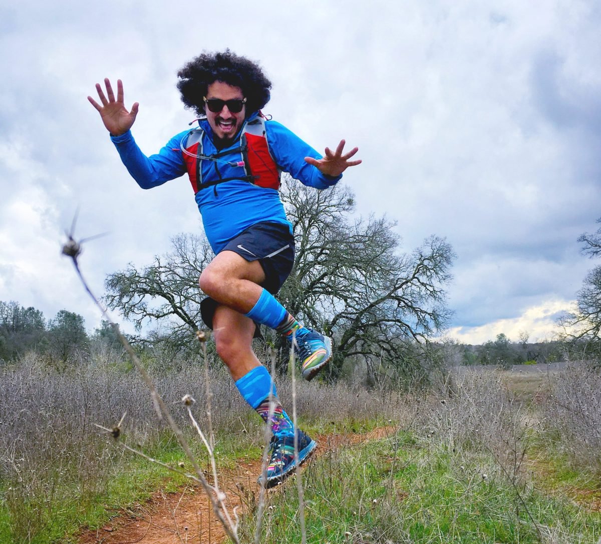 A man wearing a red running vest and blue running clothes jumping along a trail