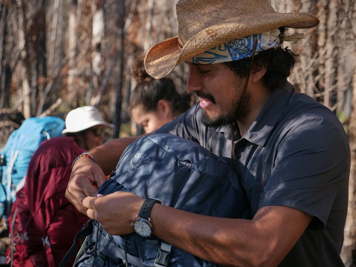 A man holding a blue backpack and wearing a tan het, with people in the background