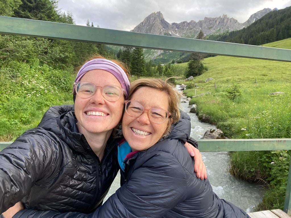 Two women wearing hiking gear, coats and glasses posing on a bridge above a river, with grass along each side and a mountain in the background