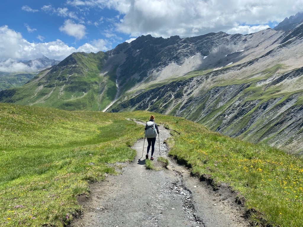 A woman hiking along a trail wearing backpacking gear and holding trekking poles, with mountains in the background