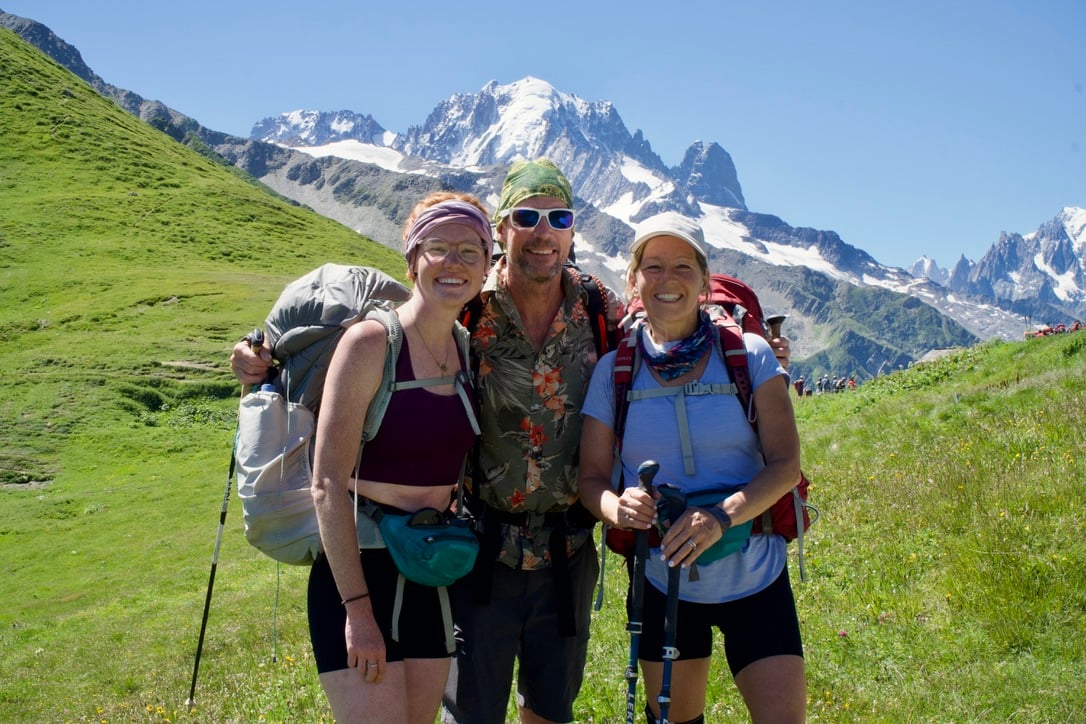 A man standing between two women, all wearing backpacking gear, with grassy fields behind them and snow-peaked mountains in the distance