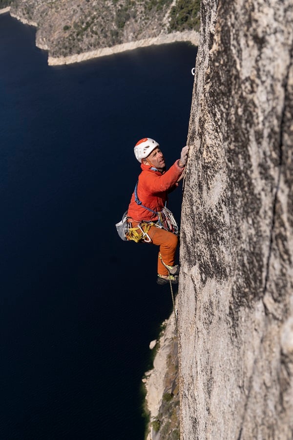 A man rock climbing up a cliff face, wearing a red top and orange pants, and a white and red helmet