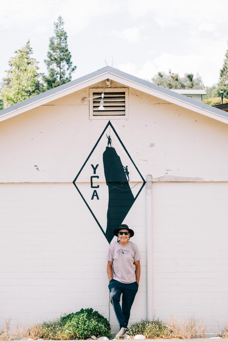 A man wearing a tan shirt, a black bucket hat and sunglasses standing in front of the YCA museum, with the YCA logo behind him