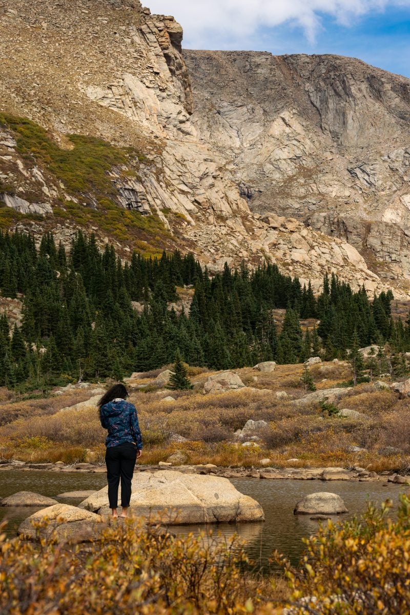 A woman standing at the edge of a lake looking off into the distance, a mountain range in the background.
