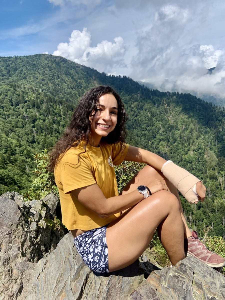 A woman wearing a yellow shirt and blue shorts sitting atop a rocky mountain overlook, with a soft cast on her left arm