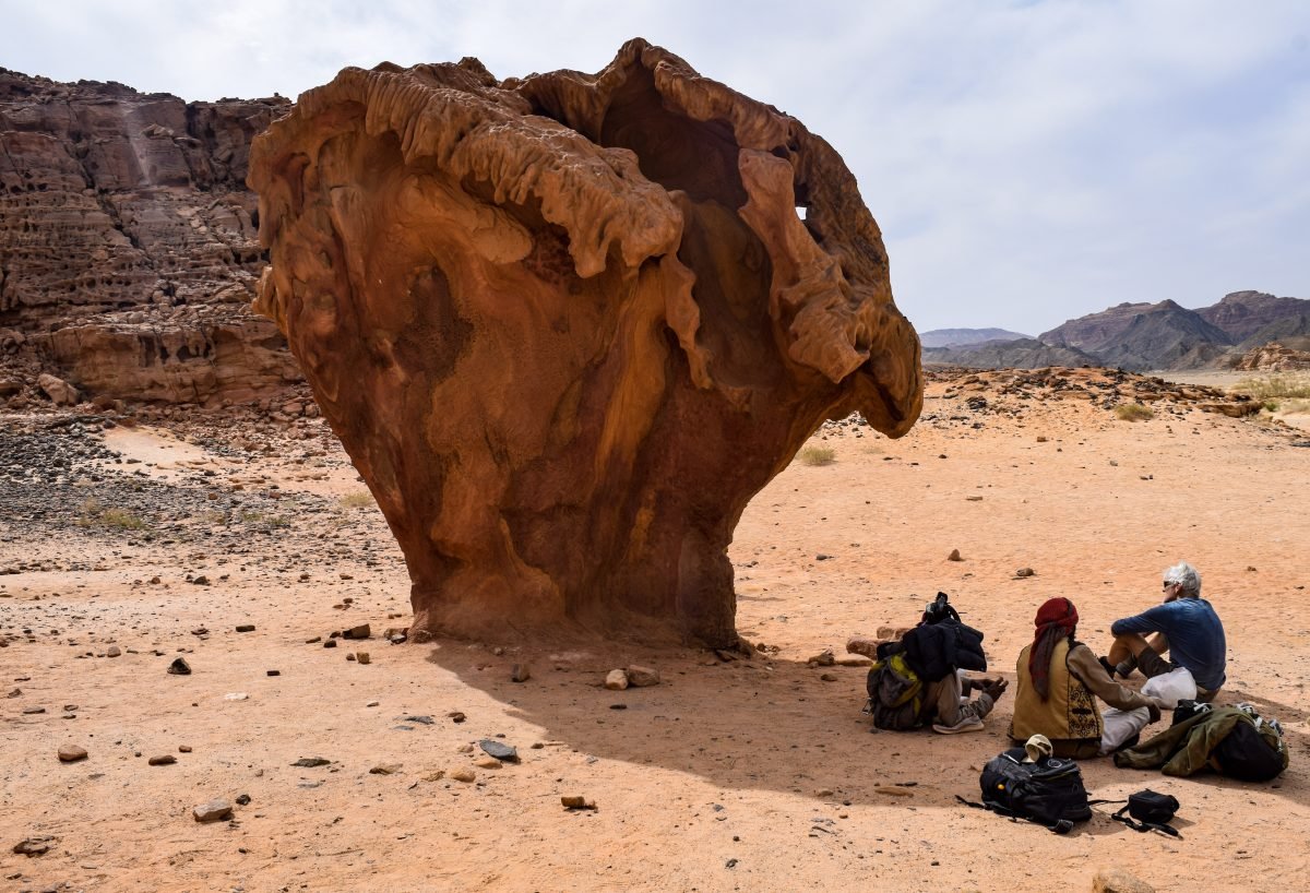 Two people sitting in the shade of a large boulder, surrounded by their backpacking gear