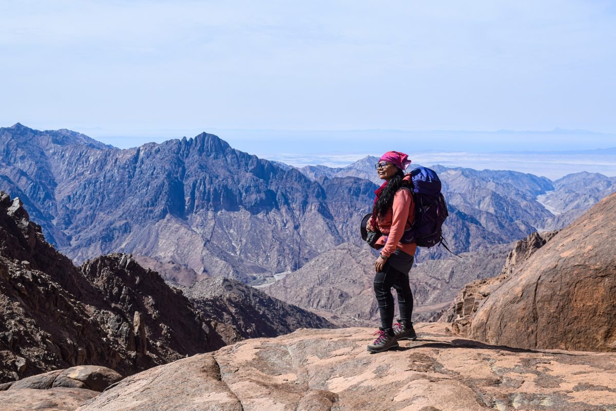 A woman in backpacking gear standing on a rocky overlook, with a mountain range in the background