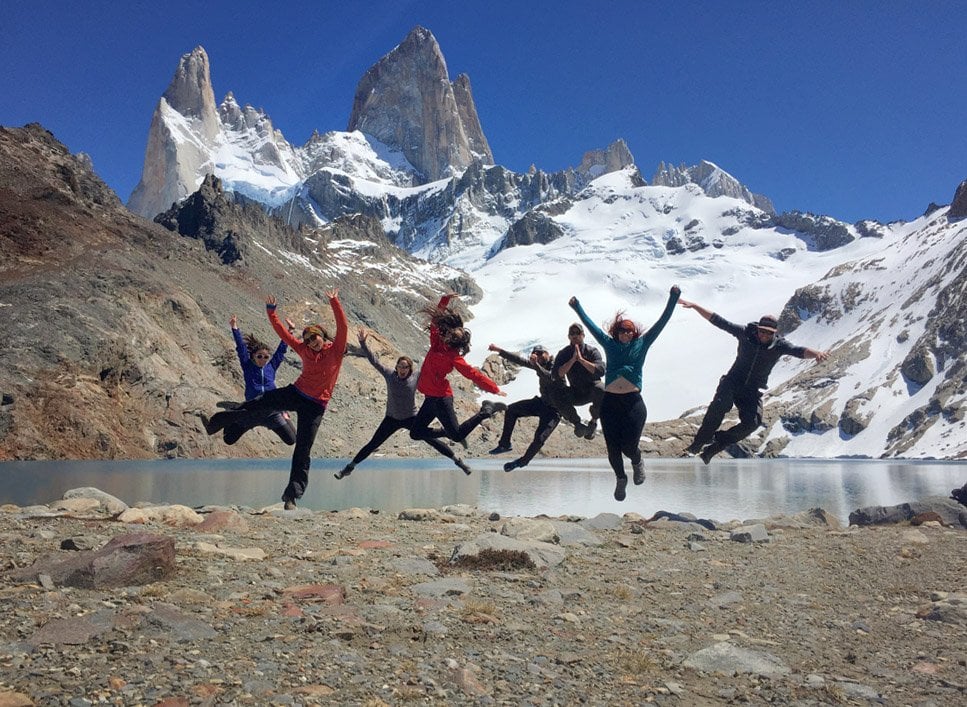 A group of people jumping and posing with an alpine lake and snowy mountains in the background