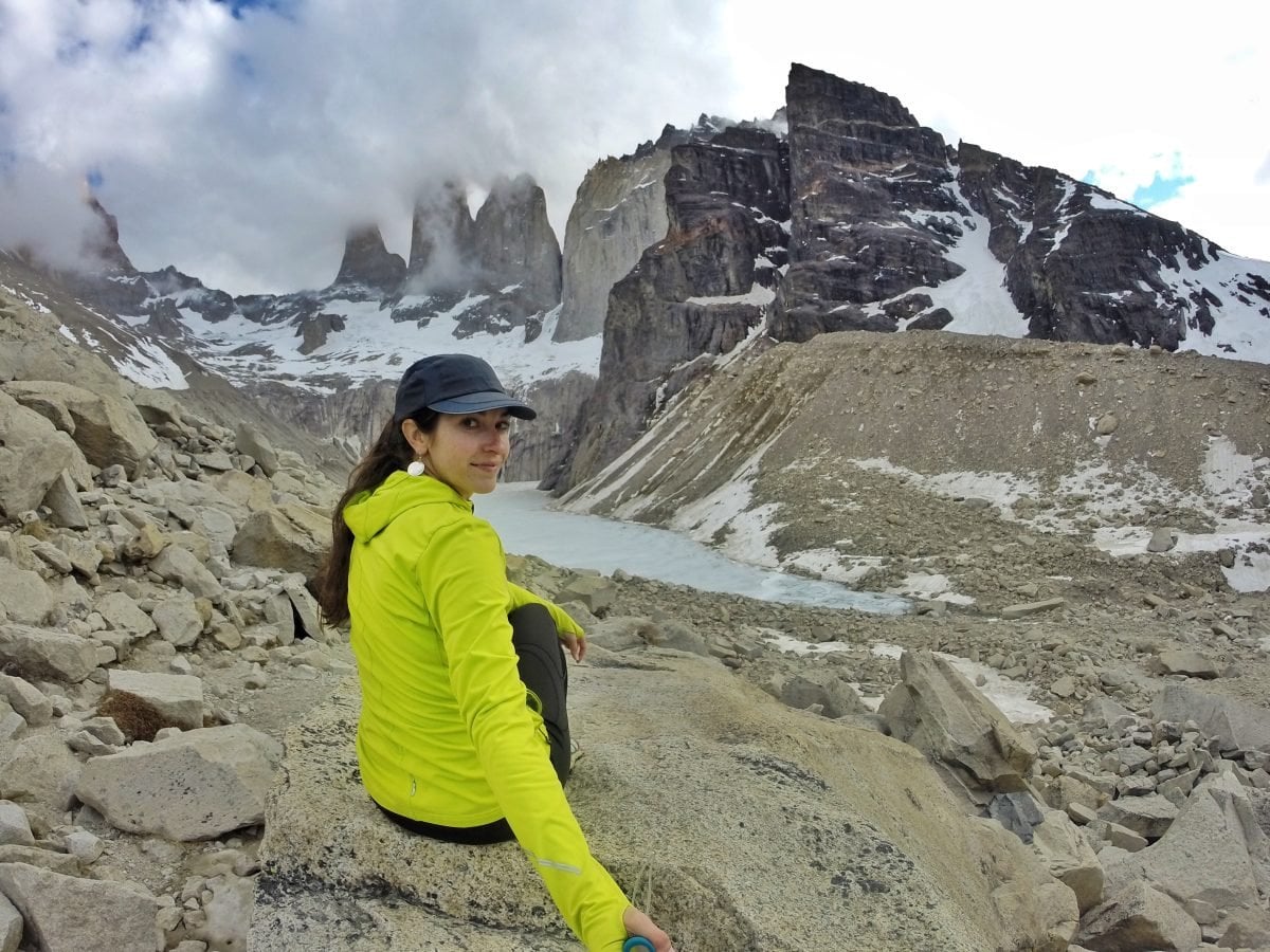 A woman wearing a bright yellow jacket and a black hat sitting on a rock that overlooks an icy alpine lake