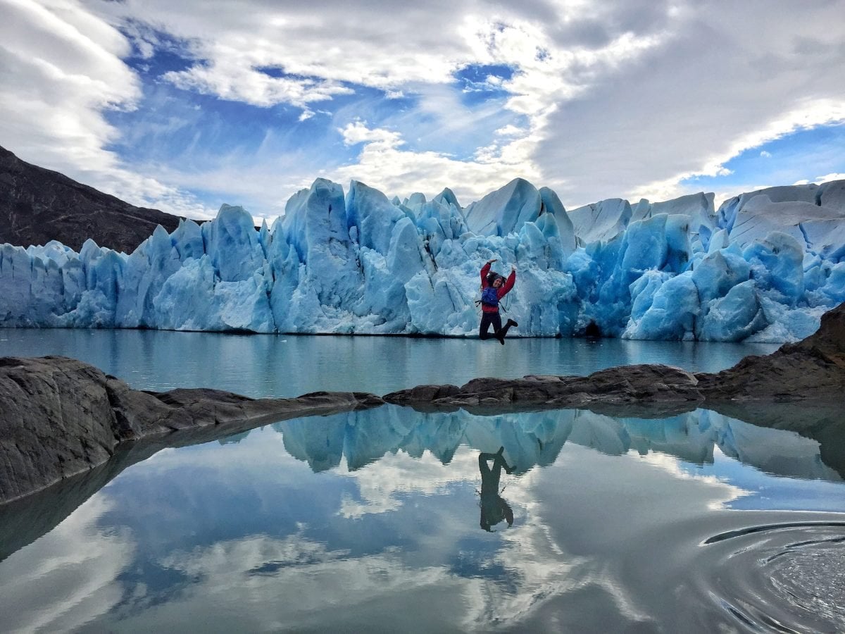 A woman wearing a red shirt and a blue life jacket jumps on a small path of rocks between two bodies of water, with glaciers in the background