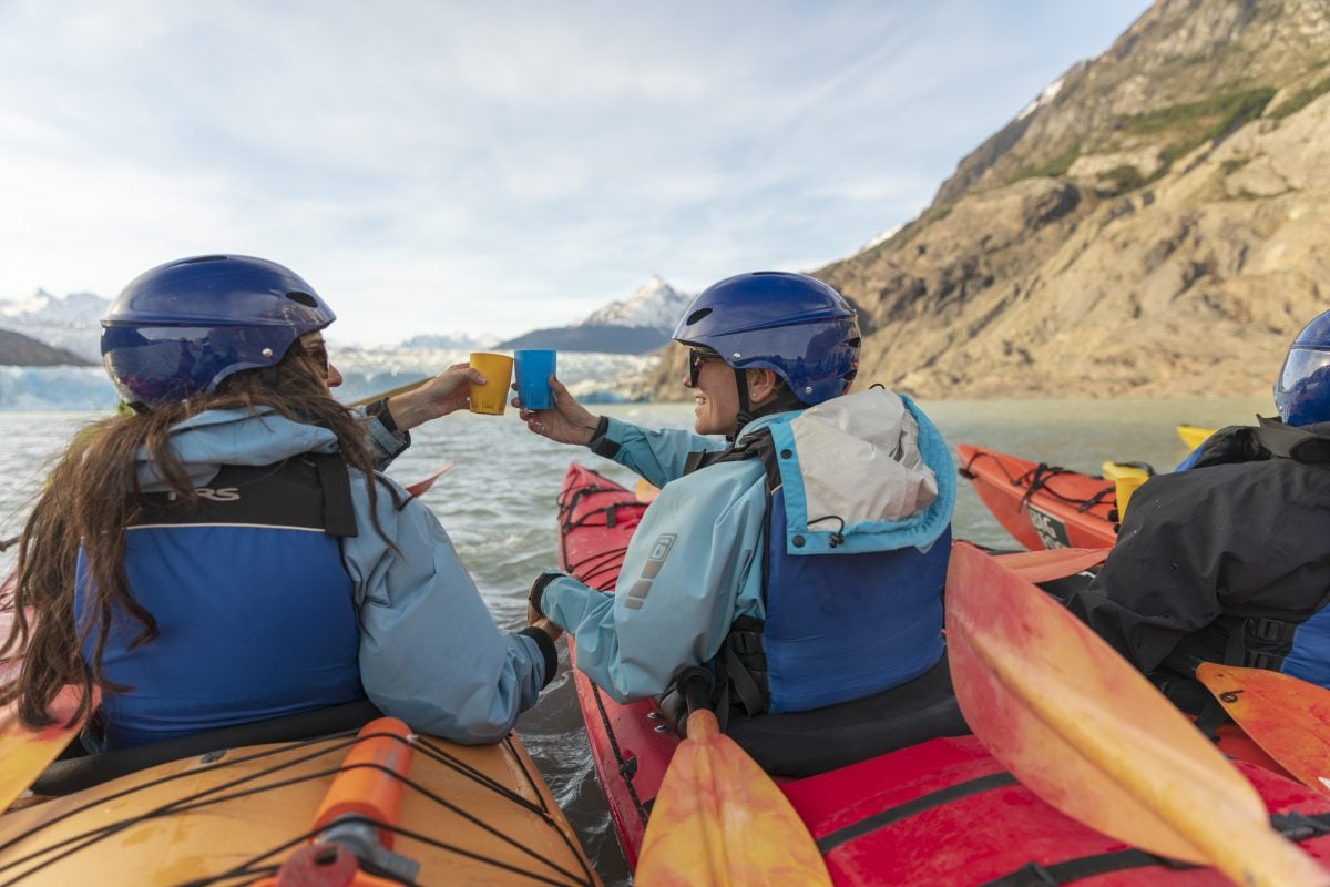Two people in kayaks wearing blue life jackets and blue helmets cheersing each other on the water