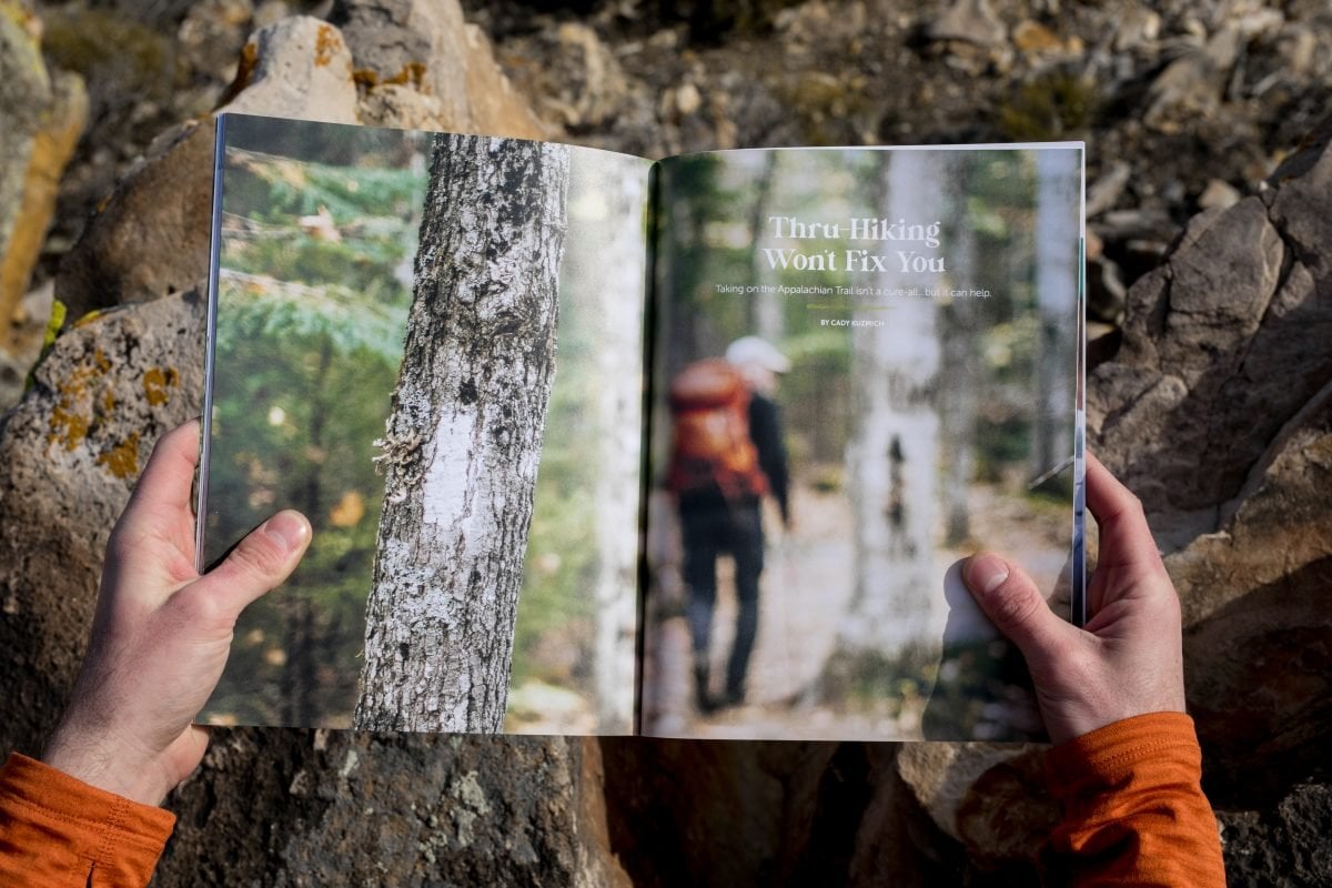 A person wearing an orange shirt holding a magazine open; inside the magazine is a feature image of a thru-hiker