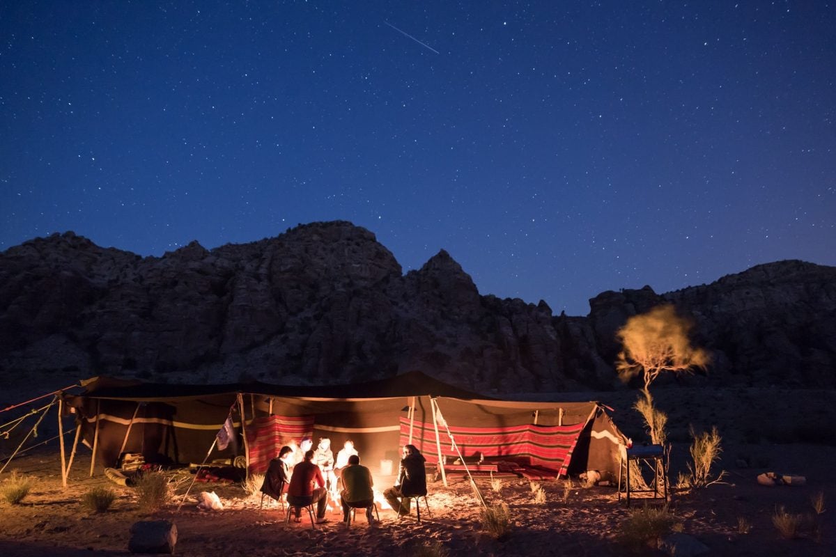 A group of people sitting by a campfire, with camping structures behind them. The night sky is lit with stars.