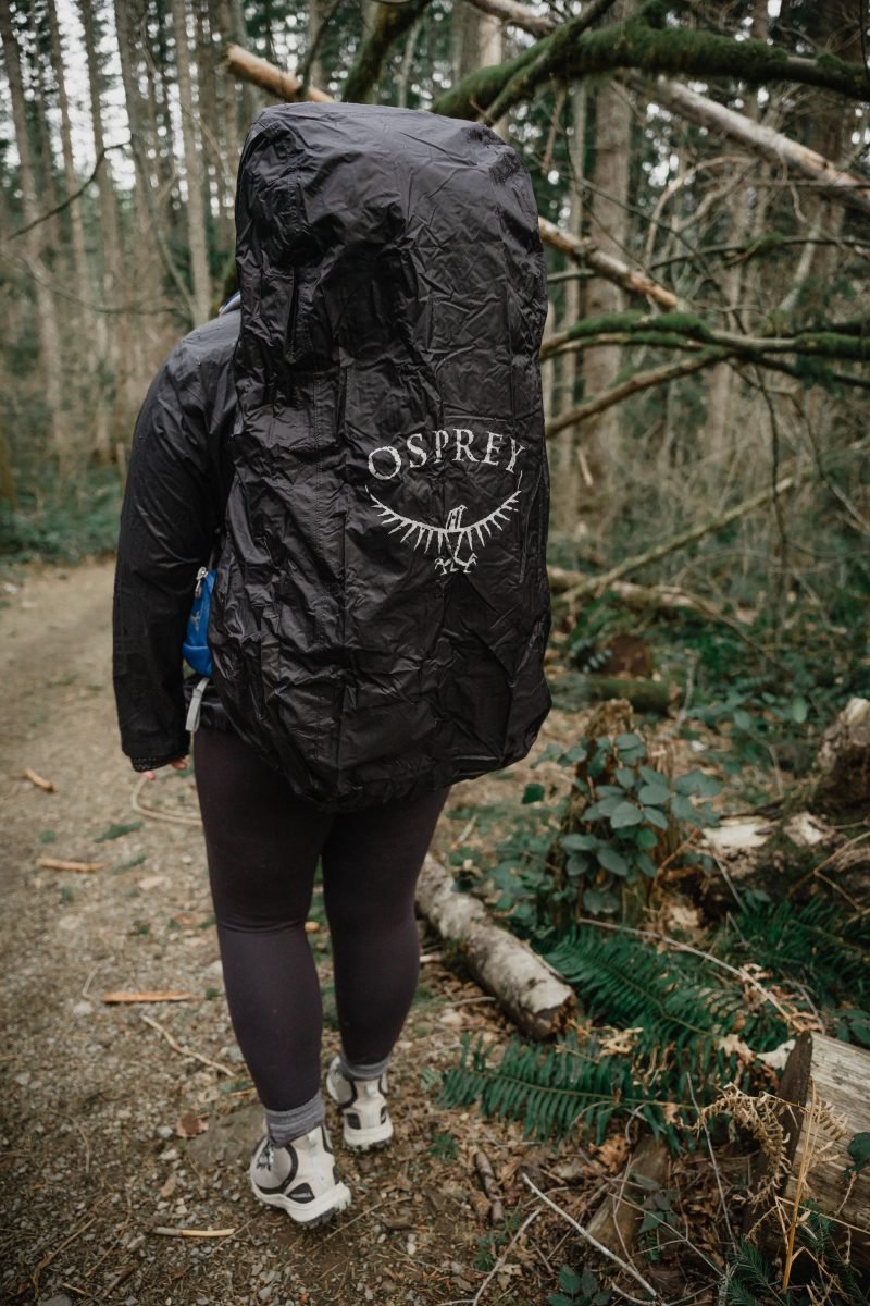A woman walking on the trail in her backpacking gear, with the black, logoed rain cover on the pack