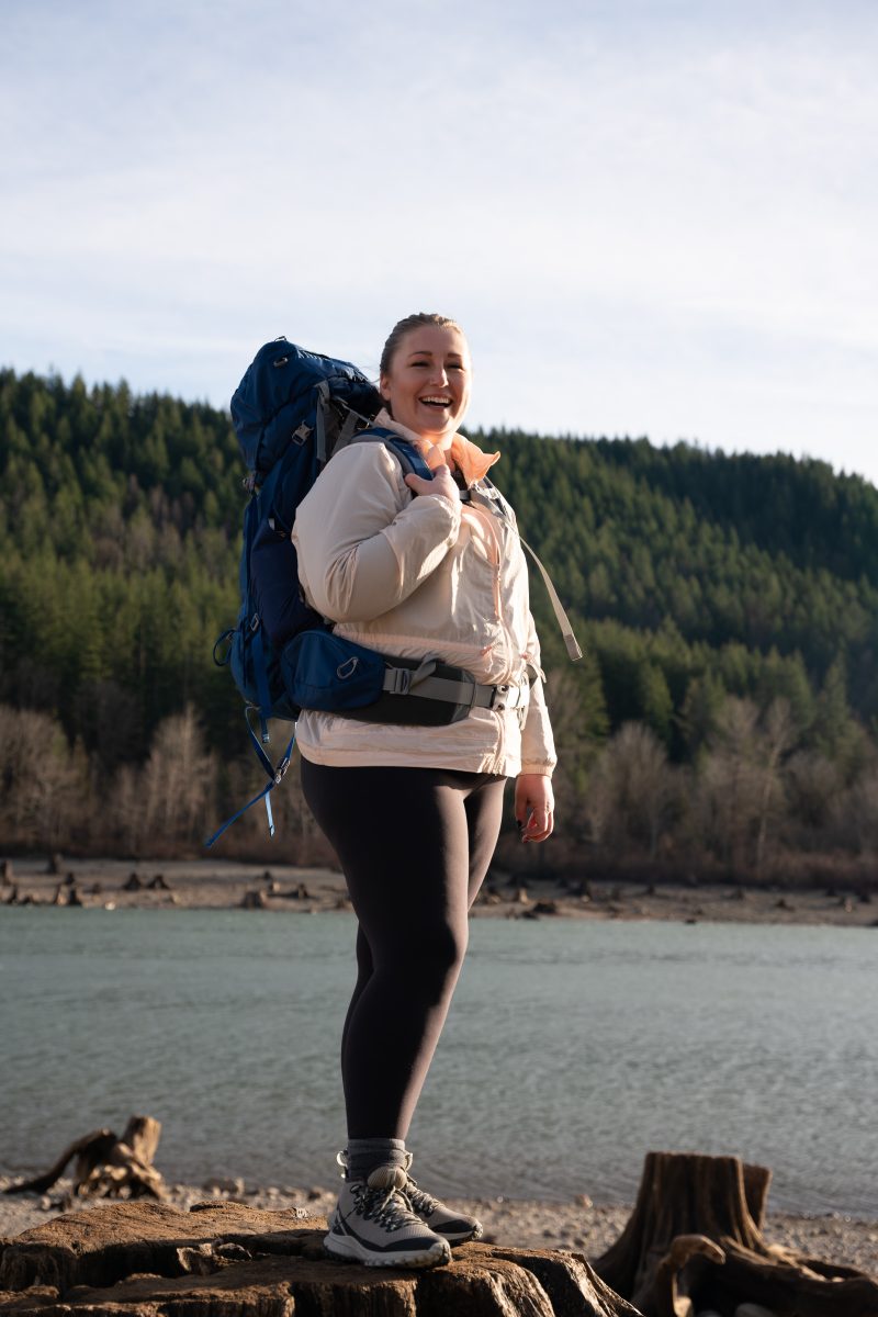 A woman standing near a body of water wearing backpacking gear.